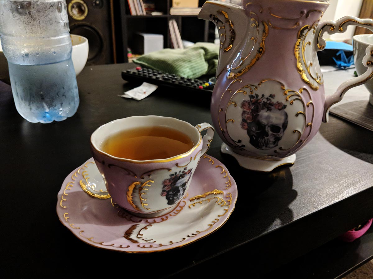 A fancy teacup in pastel pink with floral skulls, filled with tea, and a matching teapot